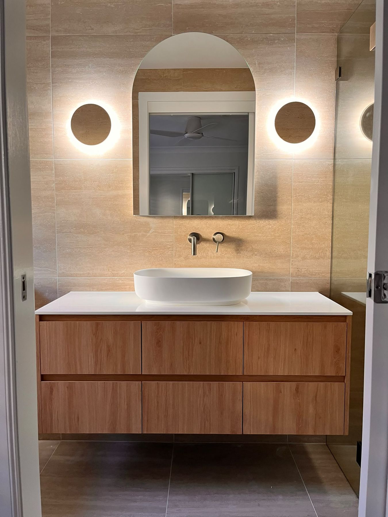 Bathroom with arched mirror, oak vanity, vessel basin and travertine look tiling